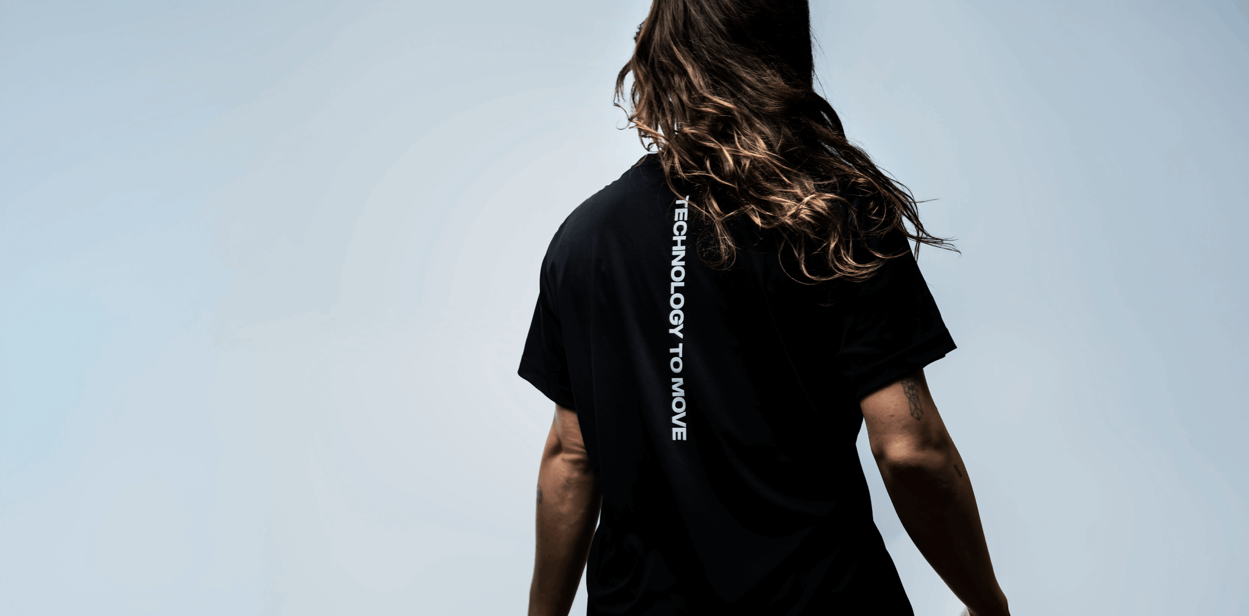 Woman with long hair with her back turned, wearing a black T-shirt which reads "technology to move".